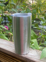 [Stainless] Silver tumbler with a clear lid attached to a wooden post with greenery in the background